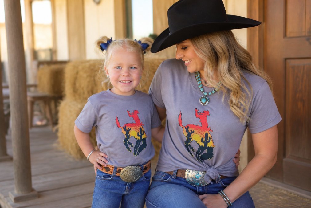 A mother and young child wearing matching western graphic tees with boots and denim, posed casually on a wooden porch with hay bales in the background, warm golden-hour lighting