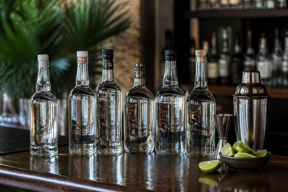 Several clear glass bottles of white rum arranged on a dark wooden bar top, backlit to show crystal clarity, with a cocktail shaker and fresh lime wedges nearby