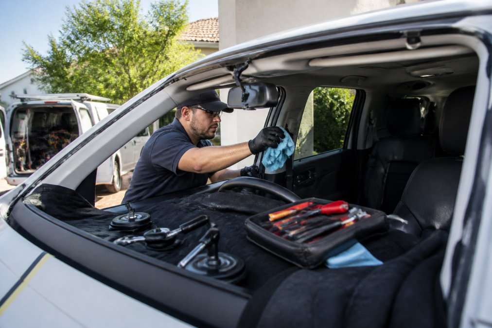 Professional technician in uniform clearing the dashboard and interior space near the windshield, preparing a vehicle for mobile replacement service, with tools and protective materials visible in natural daylight