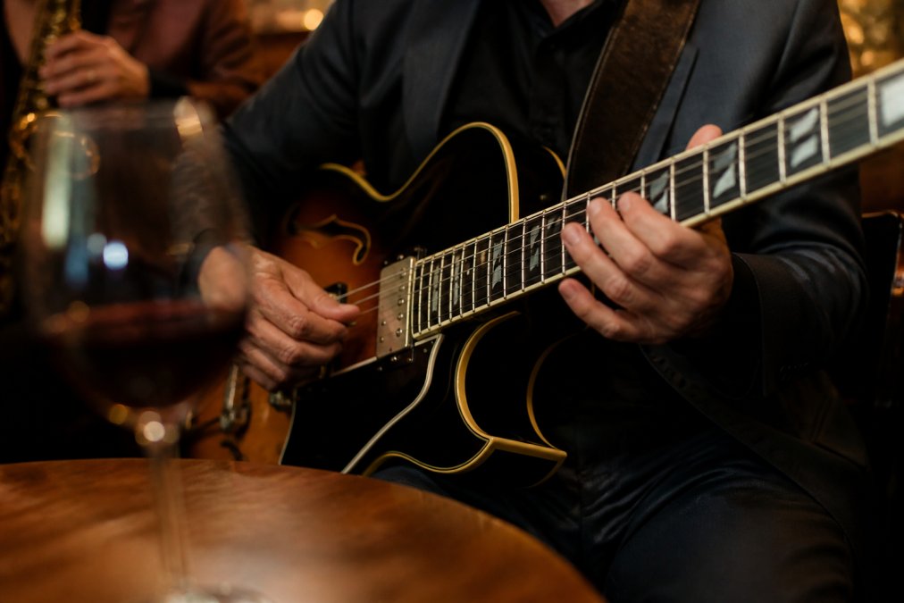 A close-up of a jazz guitarist's hands on the fretboard in a dimly lit wine bar, with a glass of red wine blurred in the foreground