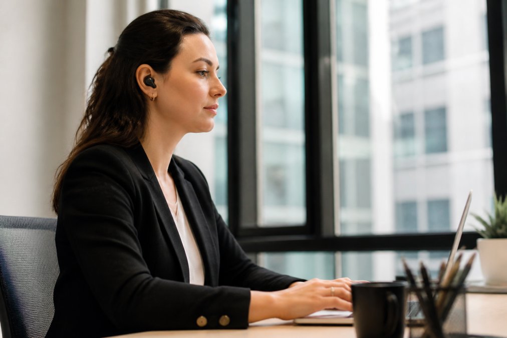 Close-up of wireless earbuds in active use during office work, highlighting connectivity and audio fidelity in a professional setting with natural window lighting