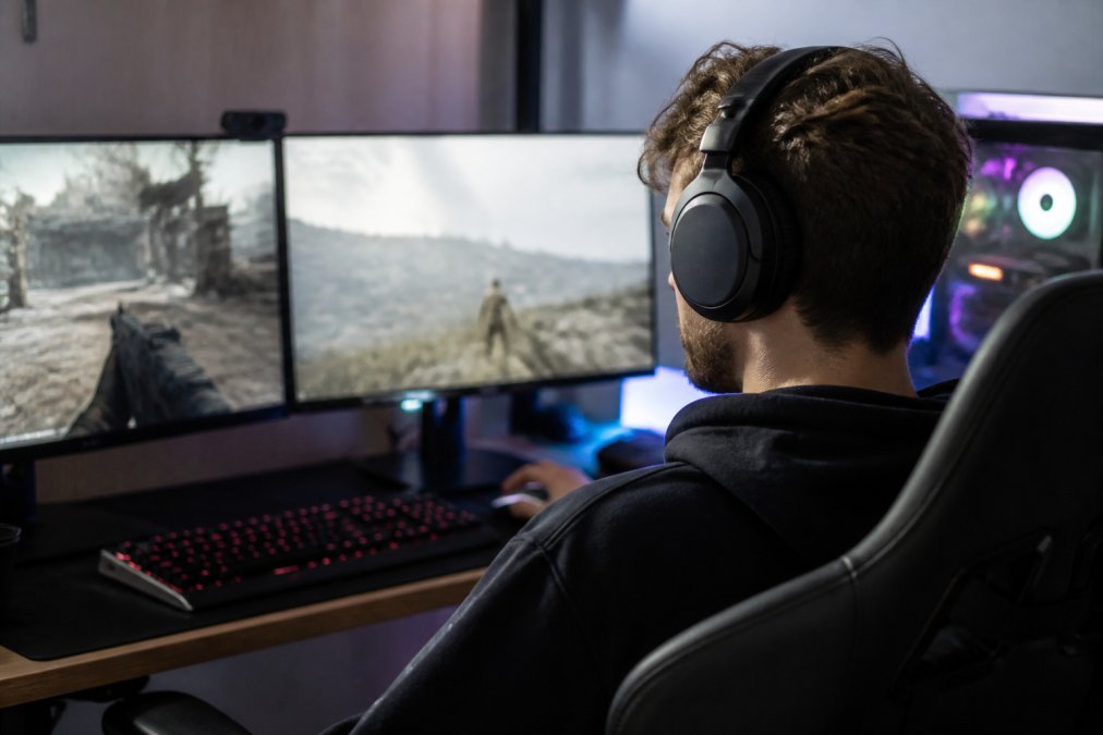 Gamer wearing wireless Bluetooth 5.3 gaming headphones at a high-end PC setup with RGB lighting, dual monitors, and gaming peripherals visible on the desk