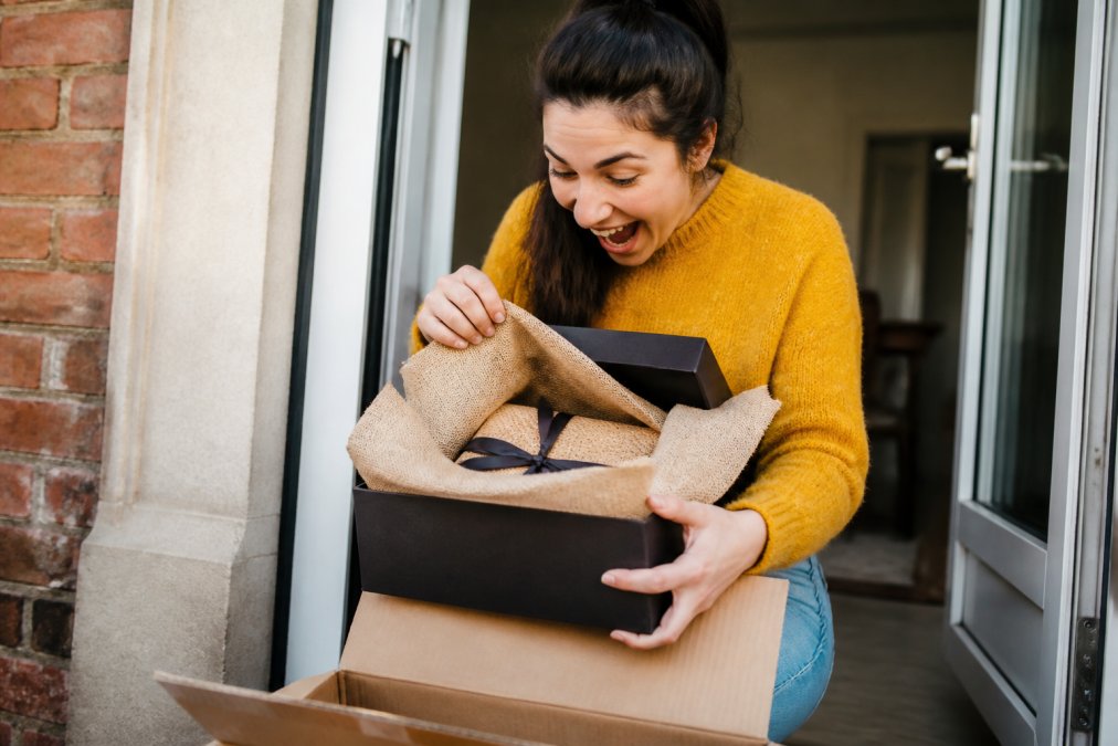 Person eagerly unboxing a carefully wrapped unique gift item at their doorstep, showing genuine excitement with the branded packaging and presentation quality