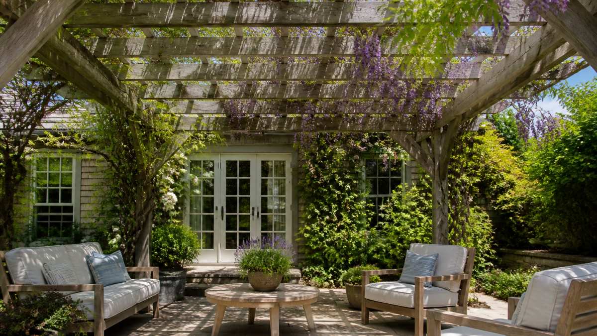 Cedar wood pergola installed over a patio with climbing vines and flowering wisteria, showing weathered but intact wood beams with natural silver-gray patina, dappled sunlight through the canopy