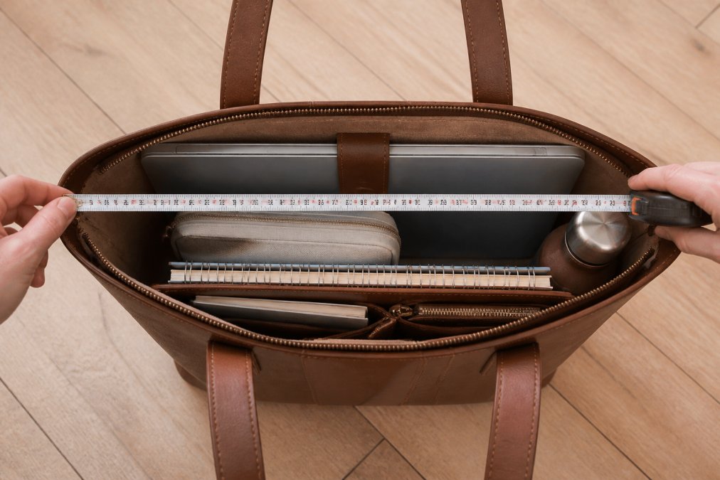 A flat-lay of a structured leather tote packed with a laptop, charger pouch, notebook, water bottle and travel card, with a tape measure showing the internal compartment width