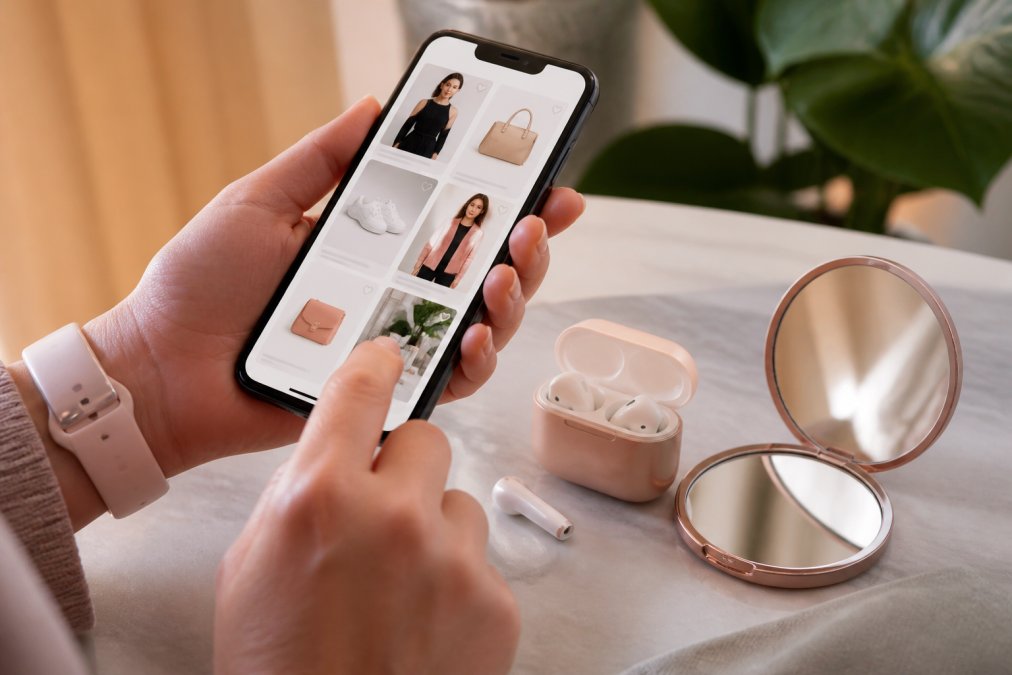 Close-up of hands holding a smartphone while browsing an online shopping app, with trendy wireless earbuds and a compact mirror visible on a desk nearby
