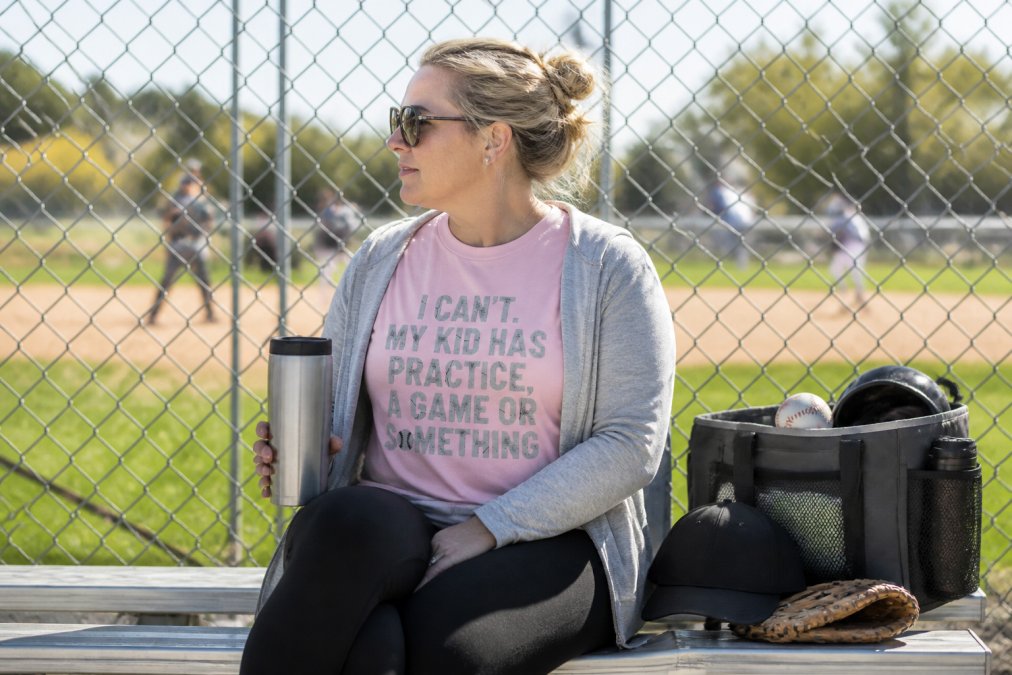 Parent sitting in bleachers at youth baseball game wearing moisture-wicking hoodie and comfortable apparel, holding travel mug with sideline gear organized nearby, natural afternoon sunlight