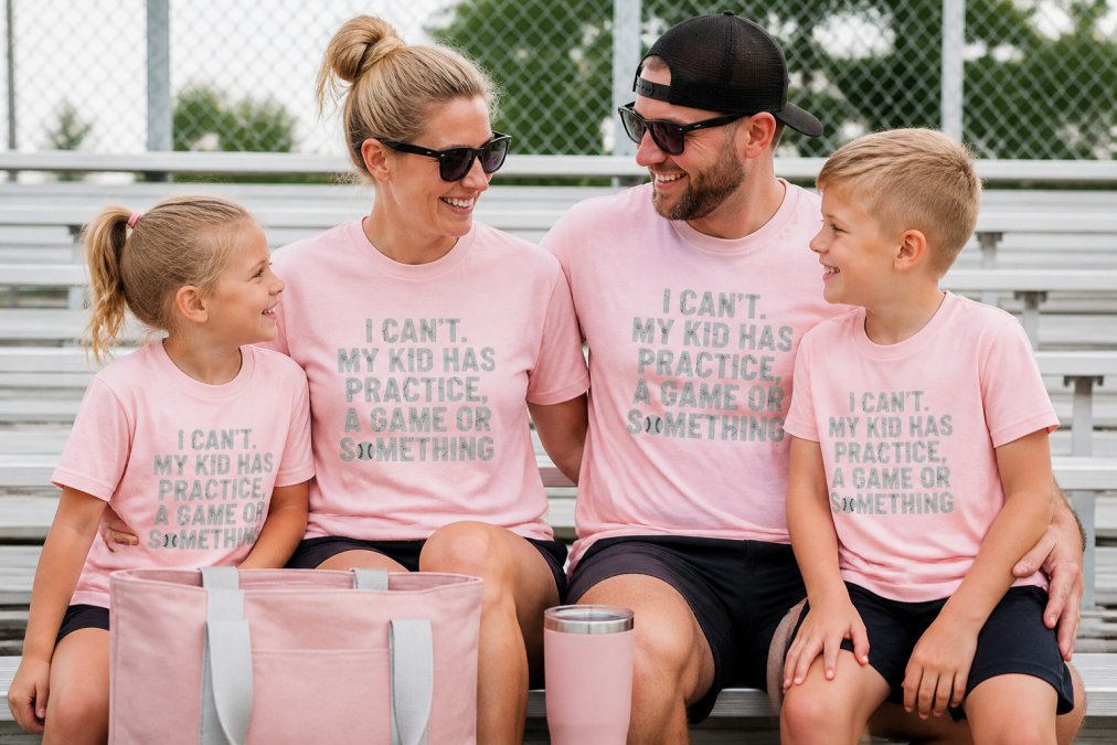 Family of four wearing coordinated custom sports team apparel at youth soccer game, sitting together in bleachers with tote bag and travel mug visible, showing complete sideline outfit coordination