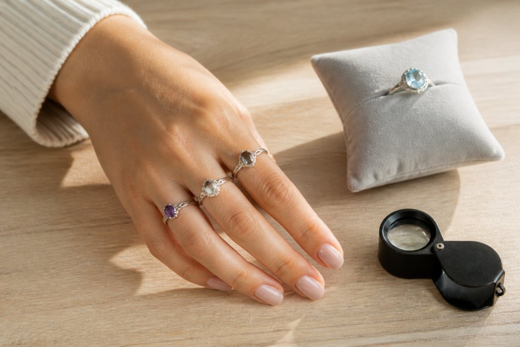 Eine elegante Hand einer Frau, die mehrere 925er Silberringe mit verschiedenen Edelsteinen an den Fingern trägt, auf einem hellen Holztisch mit weichem Tageslicht, daneben ein Samtkissen und eine kleine Lupe