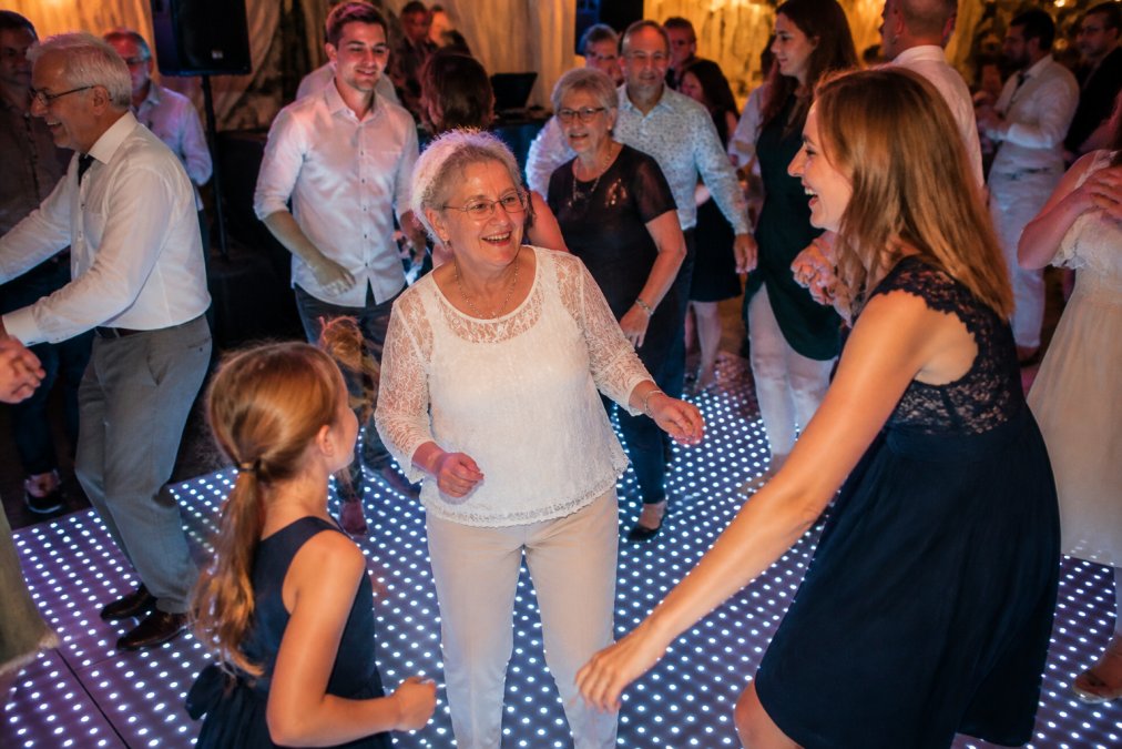 Groep mensen van verschillende leeftijden dansend op een moderne verlichte dansvloer met DJ-booth op de achtergrond, inclusief ouderen en jongeren die plezier hebben