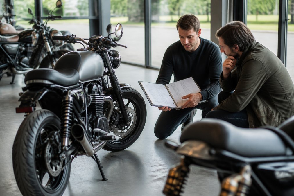 Professioneel geïnspecteerde motorfiets in moderne showroom met technicus die onderhoudshistorie-documentatie toont aan potentiële koper, natuurlijke verlichting door grote ramen