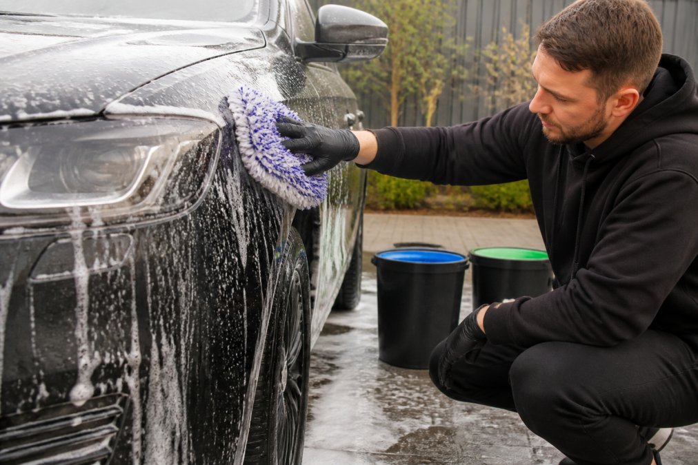 Detailer die voorzichtig een auto wast met twee emmers zichtbaar op de achtergrond, twee-emmer-systeem duidelijk, focus op krasvrijheid en zorgvuldige handeling