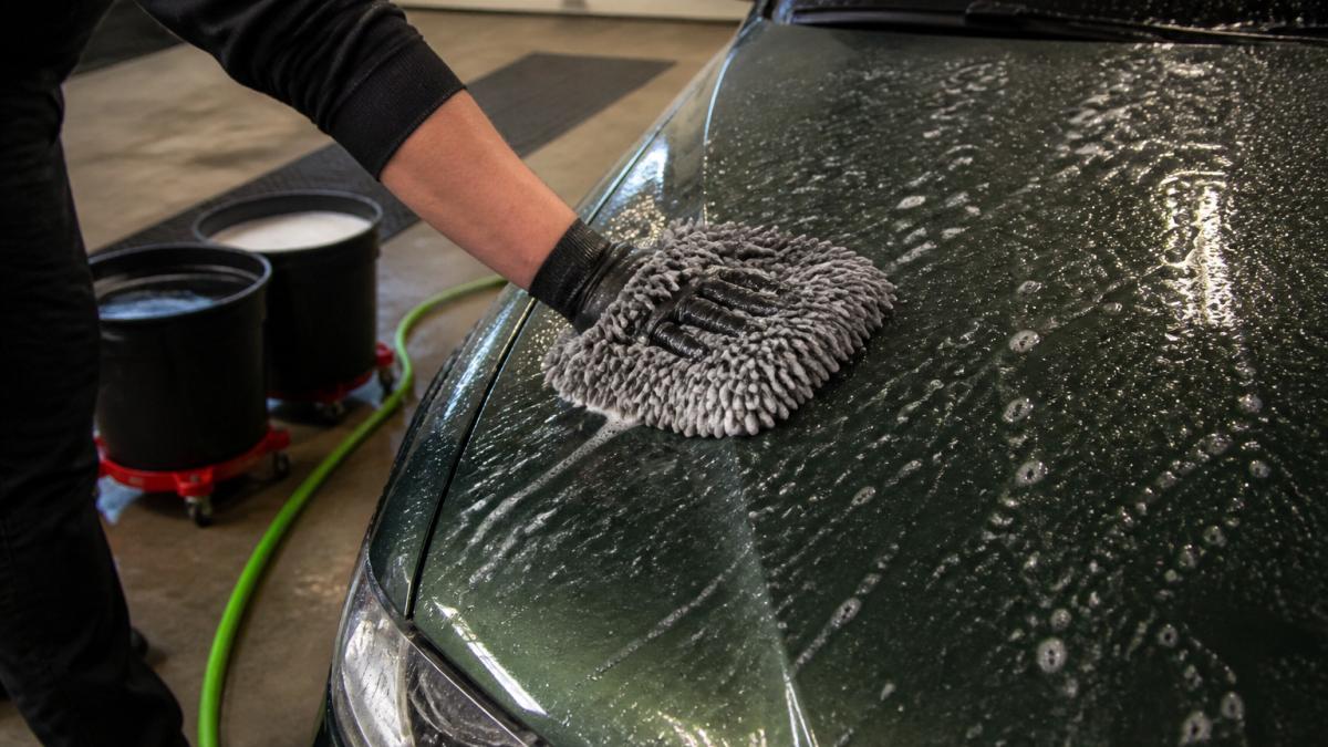 Close-up van handen in een natte washandschoen die met schuim over een donkergroene, waterafstotende autolak glijden, emmers en een tuinslang op de achtergrond in een garage