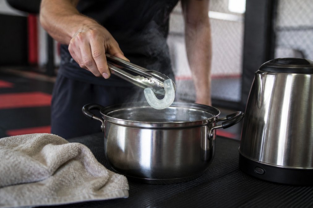 Close-up van handen die een boil-and-bite bitje met een tang uit heet water halen boven een dampende pan, met een handdoek en waterkoker op een trainingsruimte tafel