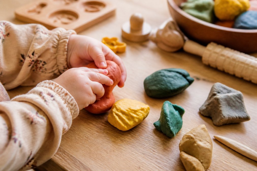 Close-up van kleine kinderhandjes knedend met gekleurde natuurlijke speelklei in verschillende bouwkleuren, met speelklei-accessoires zichtbaar op de achtergrond