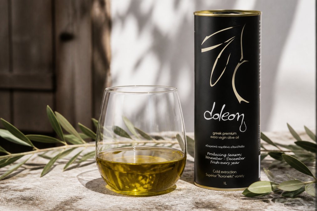 Close-up van een glas Doleon olijfolie met karakteristieke gouden-groene kleur gefotografeerd tegen natuurlijk licht, met originele verpakking zichtbaar op de achtergrond