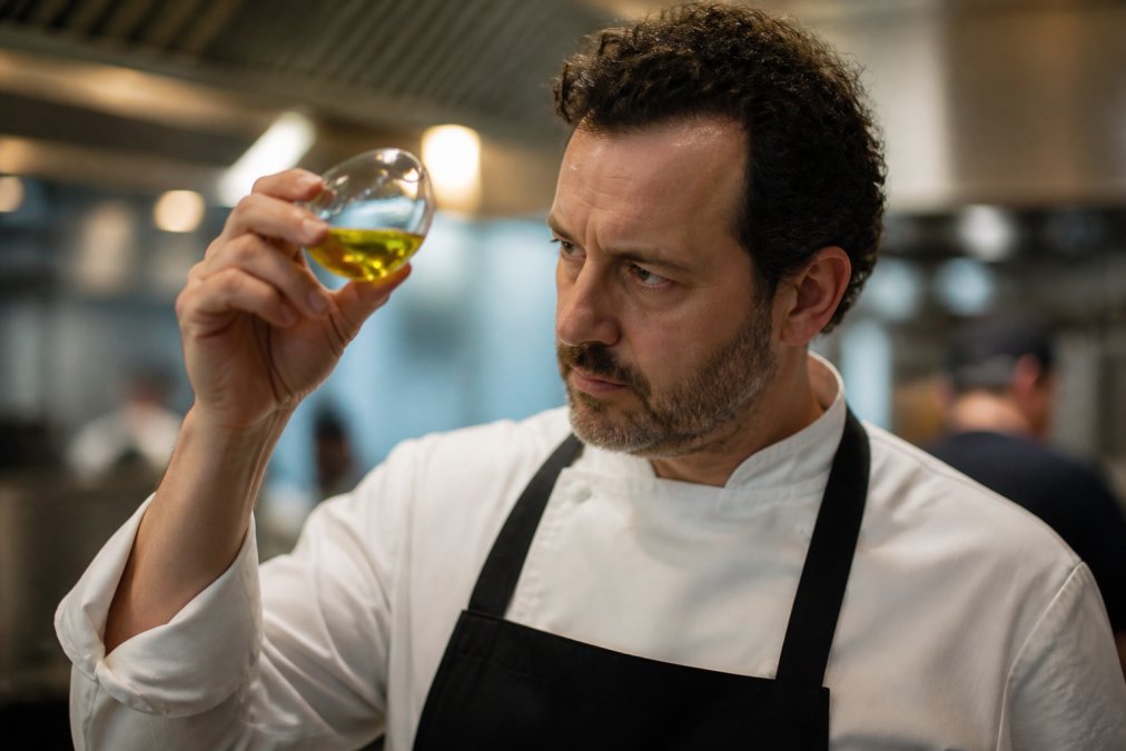 Close-up van een chef die extra vierge olijfolie uit een helder glas proeft, waarbij de kleur en helderheid van de olie tegen het licht zichtbaar zijn in een professionele keukenomgeving