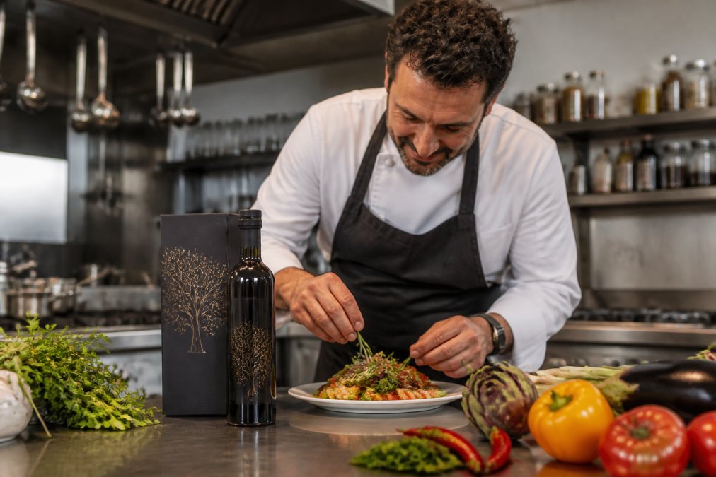 Een fles Doleon Ultra Premium extra vierge olijfolie in originele verpakking op een werkblad in een professionele keuken, omgeven door verse groenten en kruiden onder warm licht