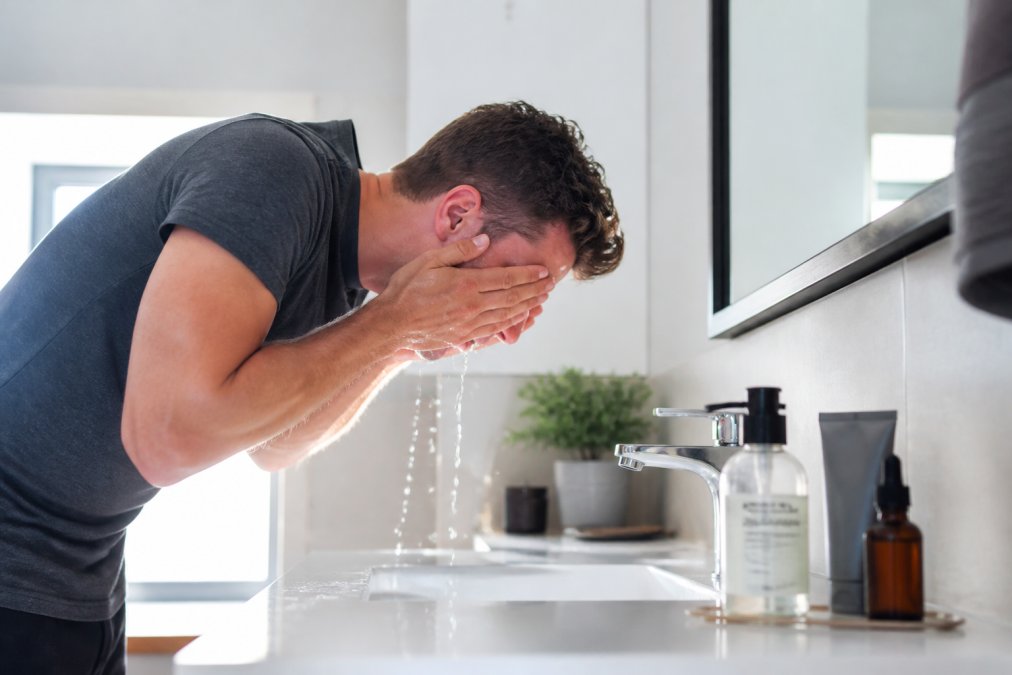 Man in badkamer die zijn gezicht reinigt met water en gezichtsreiniger, met verzorgingsproducten zichtbaar op de wastafel en in natuurlijk licht