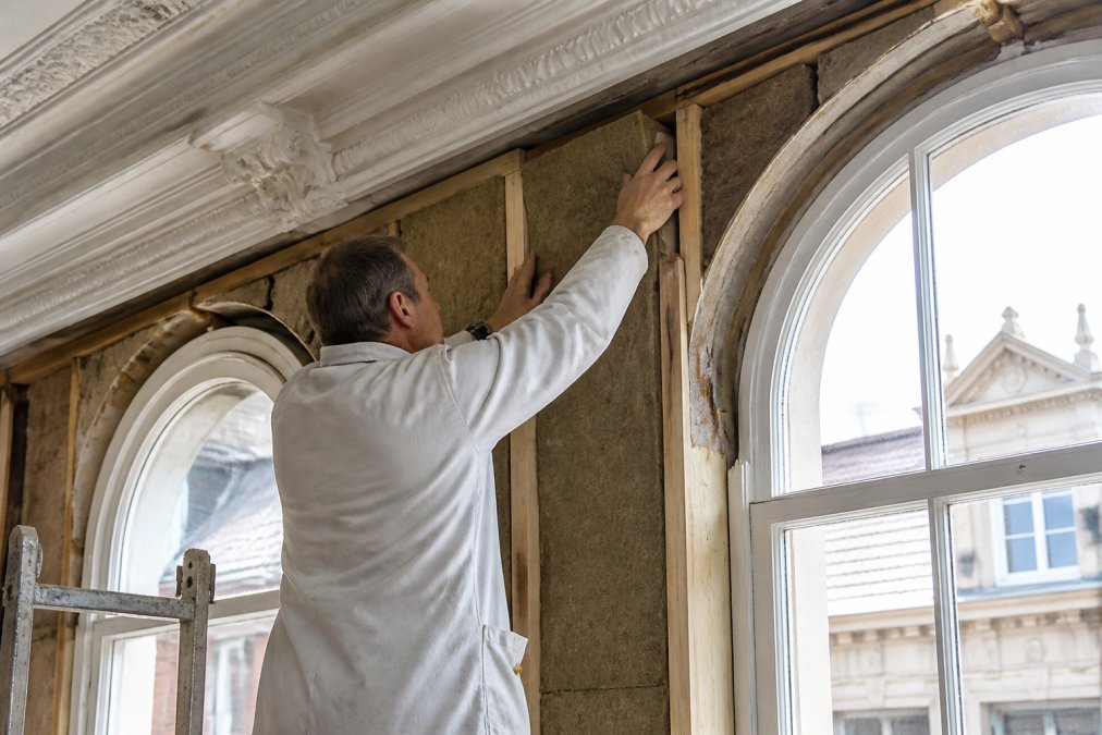 Een vakman in stofjas brengt voorzichtig dampopen isolatiemateriaal aan tegen de binnenzijde van een historische gevel, originele kroonlijst en boogramen zichtbaar, natuurlijk daglicht door het raam