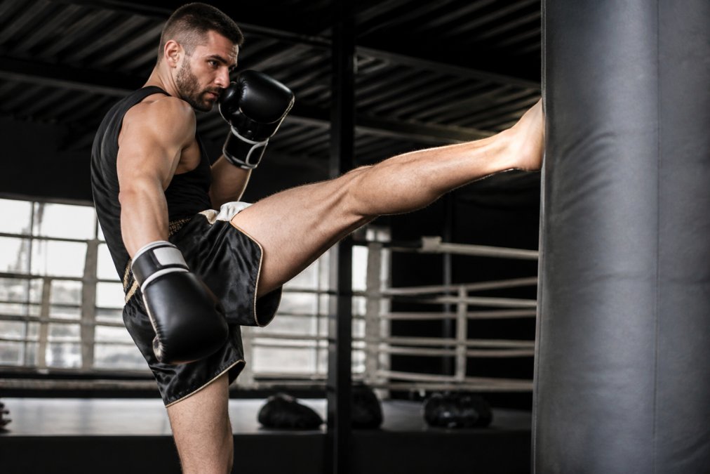 Een man in sportschoolkleding traint op een bokszak, gekleed in een satijnen Muay Thai short met zijsplit, zijn been hoog opgetrokken in een trap terwijl het broekje volledige bewegingsvrijheid toont