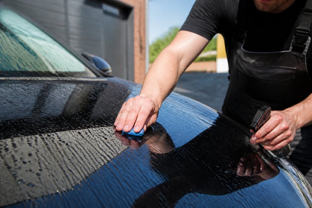 Professionele detailer die voorzichtig klei aanbrengt op glanzende autolak met glijmiddel, handen zichtbaar in werkende positie, auto in daglicht met waterdruppels
