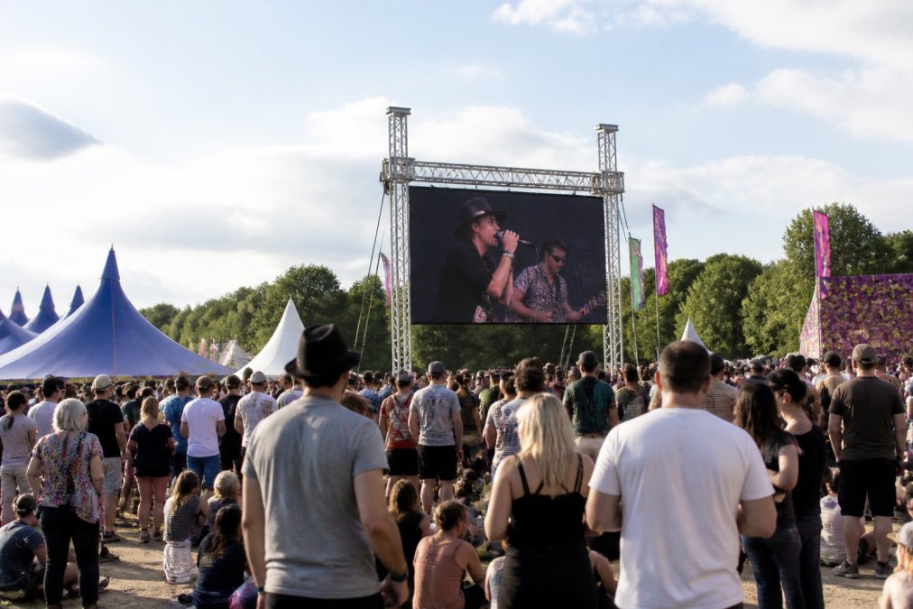 Outdoor evenement met groot LED-scherm dat live beelden uitzendt naar publiek onder daglicht, festivalsfeer met tenten, bezoekers kijken naar het scherm
