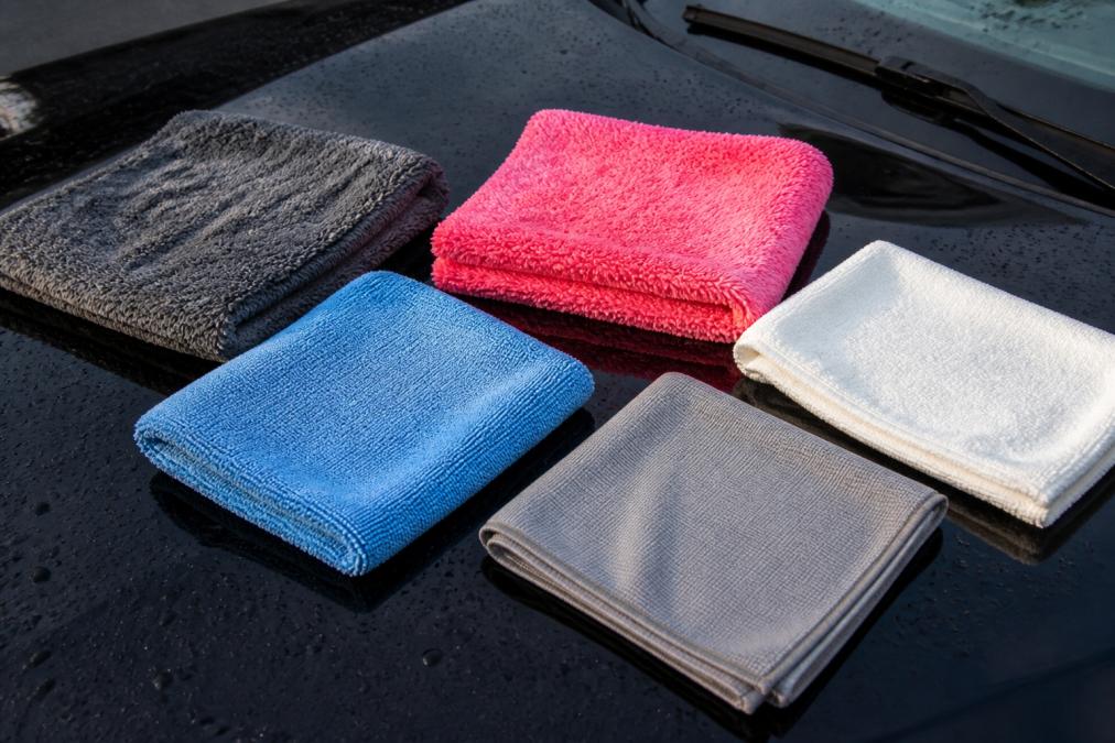 Close-up van vijf verschillende gekleurde microvezeldoeken (grijs, roze, blauw, wit) uitgespreid op een glanzende zwarte motorkap met zichtbare waterdruppels, waarbij de textuurverschillen tussen de pluche en gladde weefsels duidelijk zichtbaar zijn in daglicht