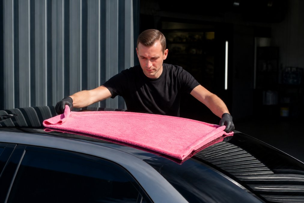 Professionele detailer die voorzichtig een grote roze droogdoek over een glanzende zwarte autodak haalt met beide handen, demonstrerend de juiste techniek met lichte druk en vloeiende bewegingen in zonnig daglicht