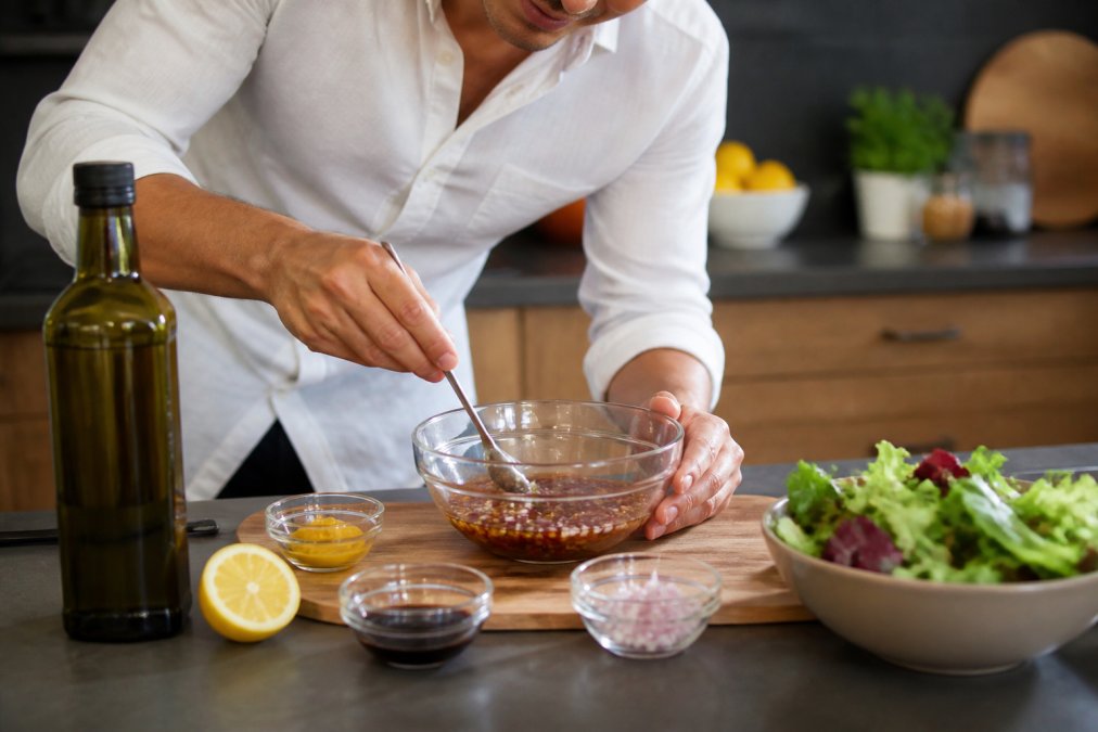 Handen die ingrediënten mengen in een helder glazen kom voor vinaigrette - olijfolie, balsamicoazijn, mosterd en fijngesneden sjalot zichtbaar, met een lepel die de mengeling emulgeert