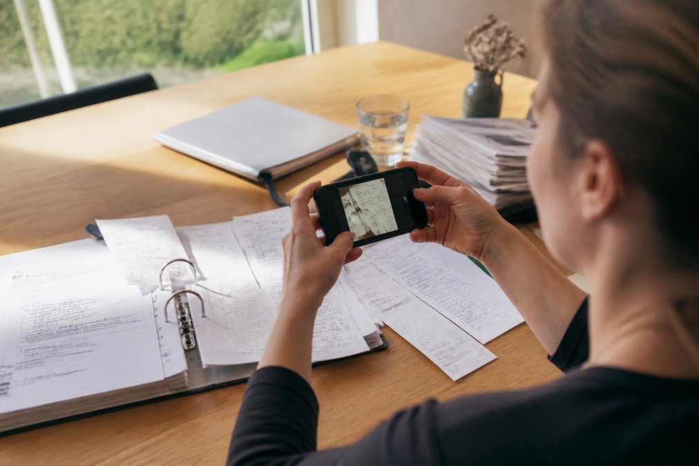 Person mit Smartphone, die Steuerrechnungen und Belege auf einem Schreibtisch fotografiert und in eine Scan-App hochlädt, umgeben von Papierquittungen und Finanzunterlagen in natürlichem Licht