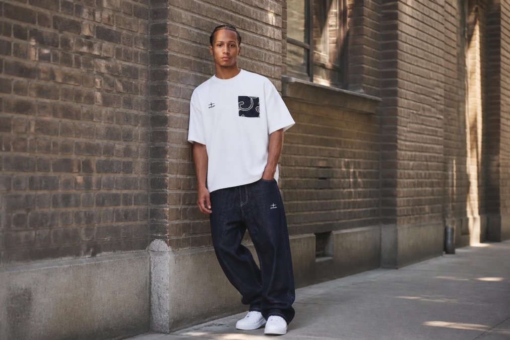 Man in oversized crèmekleurige hoodie en baggy jeans, staande in urban omgeving met minimalistisch streetwear outfit tegen bakstenen muur met natuurlijk daglicht