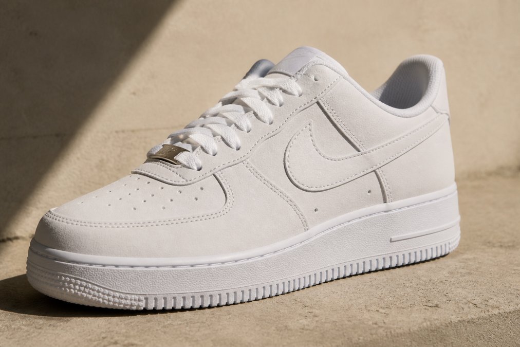 Close-up van een witte Nike Air Force 1 op een neutrale beige ondergrond, met scherpe focus op de details van de nubuck-afwerking en het klassieke swoosh-logo, natuurlijk daglicht van links