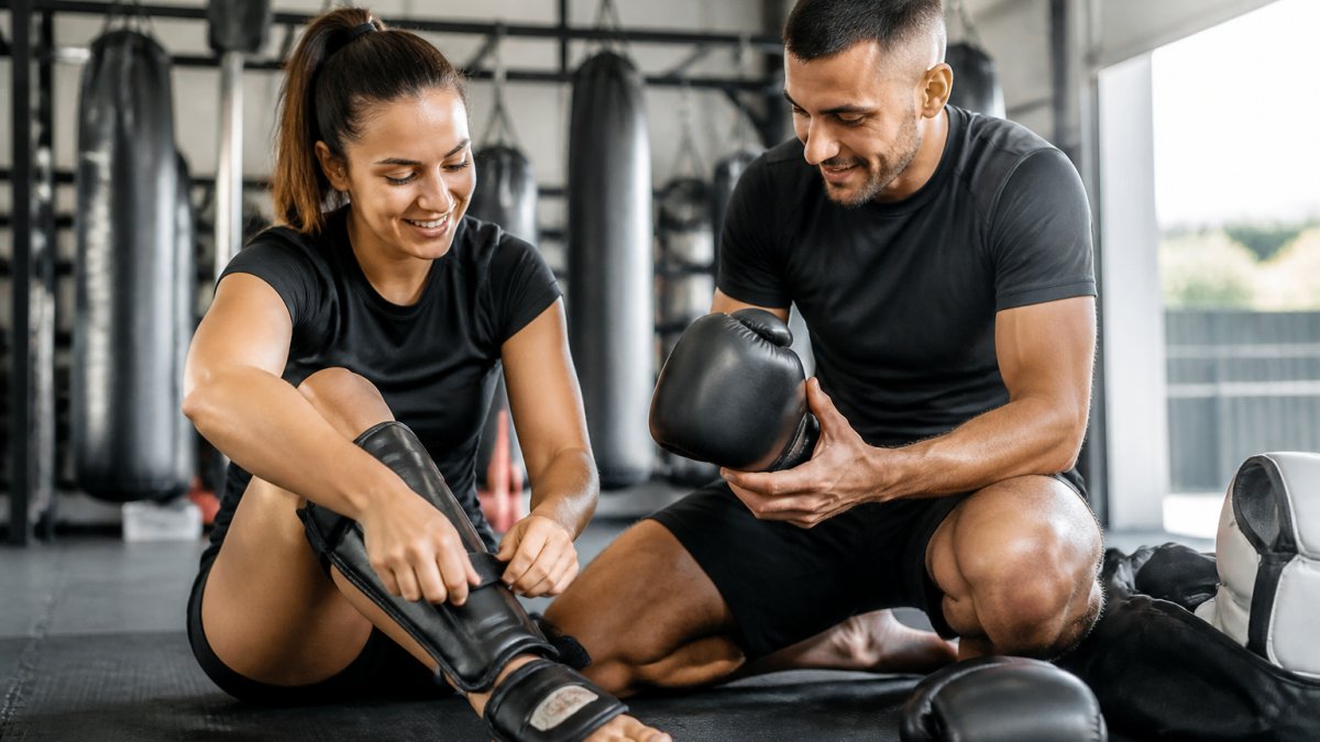 Klanten in een trainingsruimte die bokshandschoenen en scheenbeschermers controleren, met zichtbare tevredenheid terwijl ze de uitrusting aanpassen