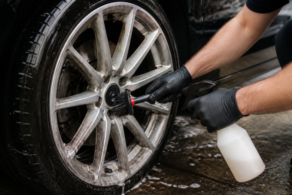 Close-up van iemand die met een velgborstel en spuitfles een lichtmetalen velg schoonmaakt, met zichtbare remstofvlekken en water dat van de velg druipt