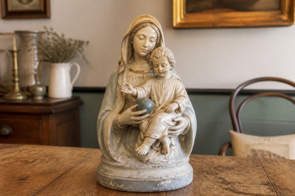 Close-up van een antiek gipsen Mariabeeld met kindje Jezus op een houten tafel in een warm verlichte woonkamer met vintage interieur, zachte kleuren en serene uitstraling