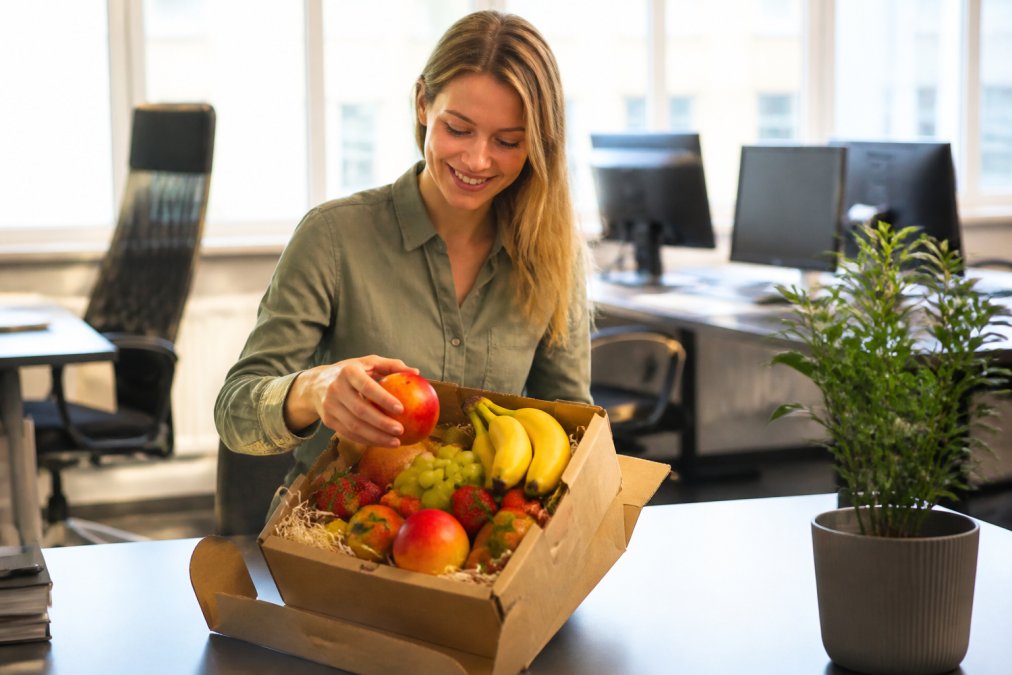 Smilende kontormedarbejder pakker en frisk frugtkasse ud ved sit skrivebord, med farverige æbler, bananer og bær synlige i kassen, i lyst naturligt kontorlys