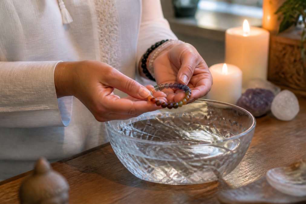 Iemands handen die een chakra armband vasthouden boven een kristallen kom met water, in een serene meditatie ruimte met kaarsjes op de achtergrond