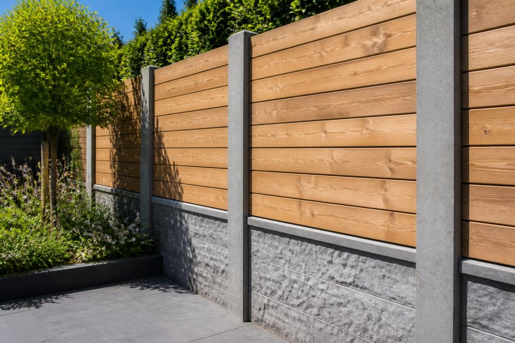 Close-up van een afgewerkte hout-beton schuttingsectie in een woontuinomgeving, met grijze betonnen palen en natuurlijke houten planken die een warm contrast vormen, zonnig daglicht