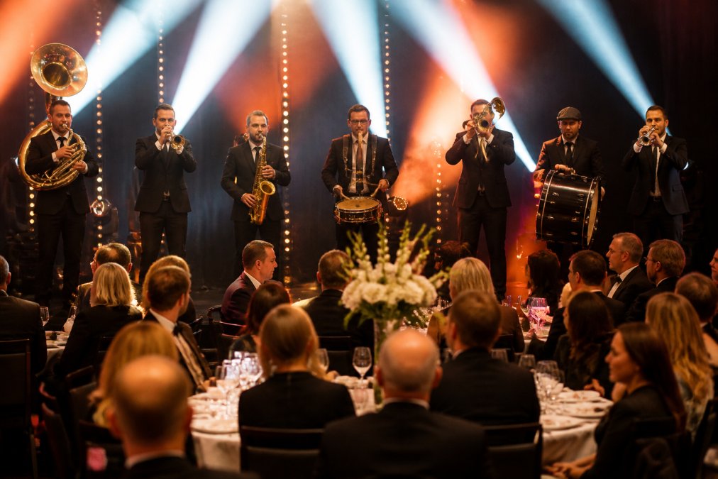 Muzikanten van een brassband in strakke gala-outfits die optreden op een verlicht podium tijdens een zakelijk galadiner, met kleurrijke lichtshow op de achtergrond en geklede gasten aan tafels op de voorgrond