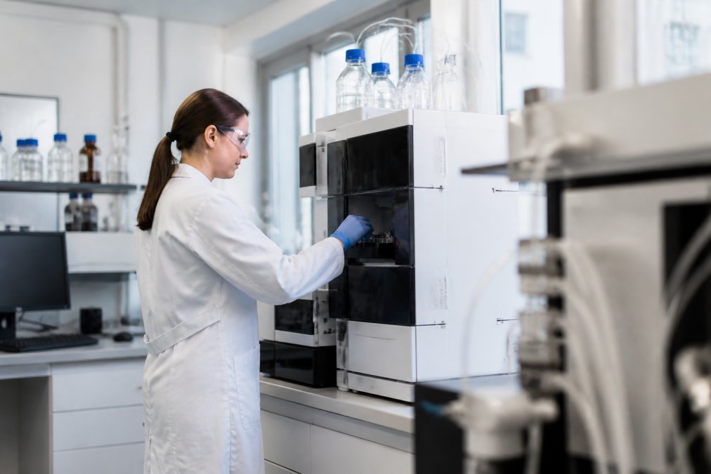 Onderzoeker in laboratoriumjas die aan een HPLC-apparaat werkt, met computermonitor die analytische gegevens en chromatogrammen toont, in een moderne onderzoekslaboratoriumomgeving met natuurlijk licht