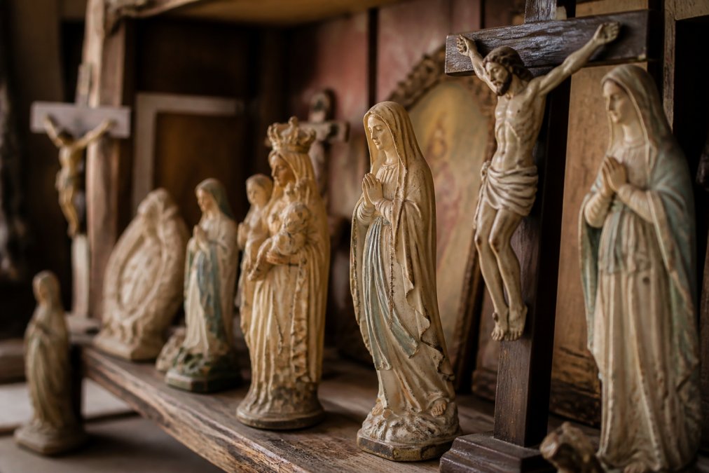 Close-up van antieke religieuze beelden op een houten plank, inclusief Mariabeelden en kruisbeelden met warme, zachte verlichting die de patina en details benadrukt