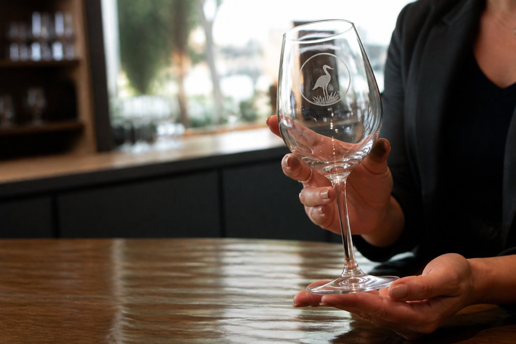 Close-up of hands holding a custom-etched wine glass with a company logo, set on a polished wooden table in a bright showroom, soft natural light catching the engraving