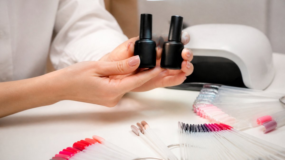 Close-up of a nail technician's hands holding two professional gel polish bottles side by side under a salon lamp, color swatches fanned on the desk