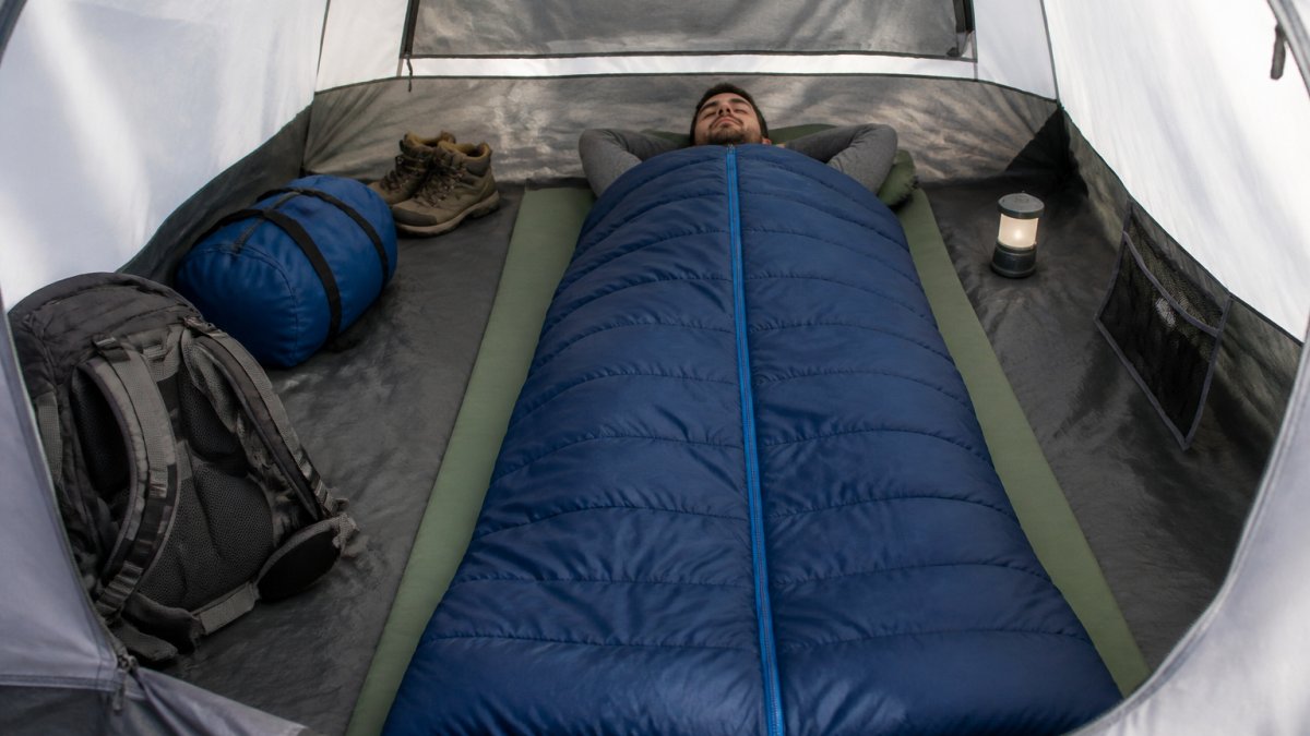 A tall camper lying inside a spacious rectangular sleeping bag in a tent, showing visible room at the head and feet, with camping gear nearby in soft daylight