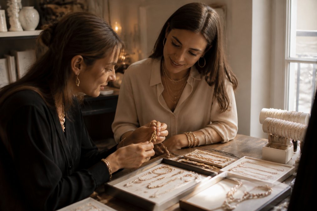Plan rapproché d'un collier artisanal en perles et fleurs faites main posé sur un tissu en lin clair, à côté d'une boîte cadeau élégante, lumière naturelle douce d'après-midi