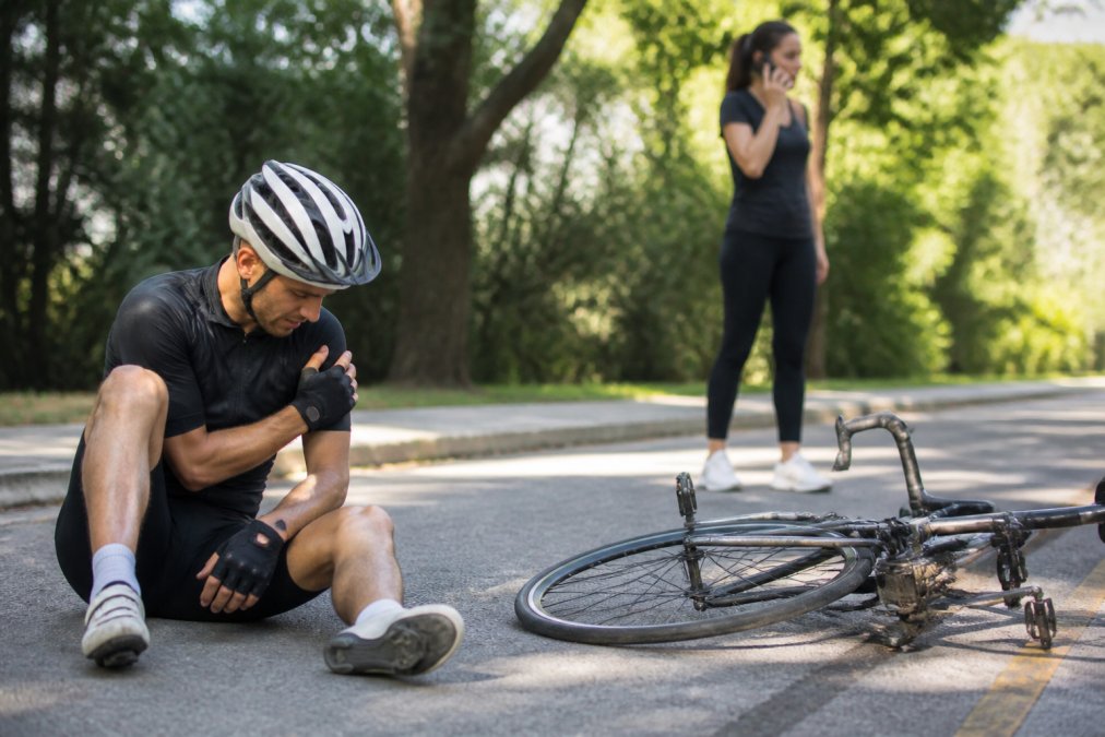 A cyclist sitting on the roadside after an accident, with a damaged bicycle nearby and another person calling emergency services on their phone, natural daylight