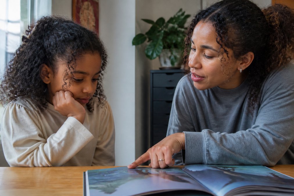 En voksen og et barn sitter ansikt til ansikt med en åpen bildebok mellom seg, den voksne peker på illustrasjoner mens barnet ser tankefullt ut og engasjert