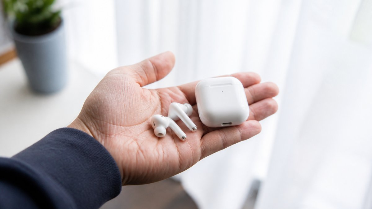 Close-up of a person's hand holding white wireless earbuds with charging case visible, minimalist background with soft natural lighting