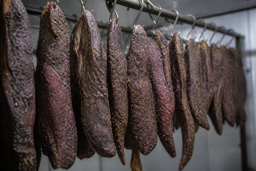 Close-up of strips of biltong hanging vertically in a temperature-controlled drying room, showing deep mahogany coloring and natural texture, with soft diffused lighting highlighting the cured meat's surface